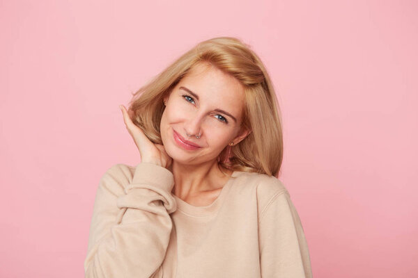 Indoor shot of young woman flrting with boyfriend, feels shy, smiles and touching her hair. Wears yellow sweater. Isolated over pink backdrop.