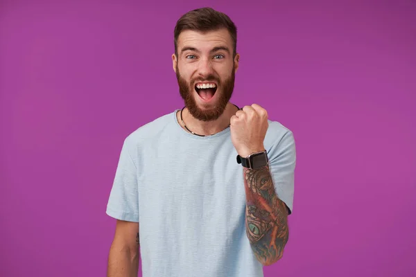 Happy handsome young tattooed brunette man with beard keeping raised fist and shouting cheerfully while looking to camera, posing over purple background in casual clothes — Stock Photo, Image