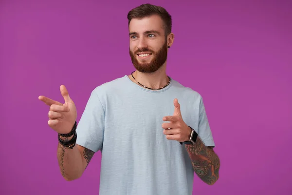 Positive pretty tattooed brunette guy with trendy haircut wearing blue t-shirt, pointing aside with forefingers and smiling widely, posing over purple background — Stock Photo, Image