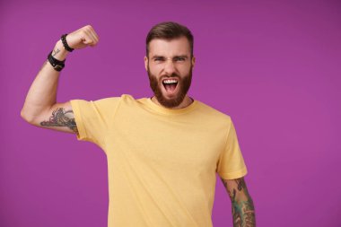 Heyecanlı, sakallı esmer, dövmeleri olan, elini kaldıran ve gücünü gösteren, ağzı açık, mor arka planda poz veren, sarı tişörtlü bir adam.