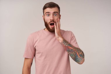 Şaşkın, genç, tıraşsız, esmer, kısa saçlı, avucunu yanağında tutan ve ağzını şaşırtıcı bir şekilde açan, yuvarlak gözlü ve beyaz arka planda poz verirken kaşlarını kaldıran adam.