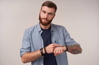Stüdyo fotoğrafında, sakallı, mavi tişörtlü ve modaya uygun aksesuarları olan, saatini gösteren ve zamanı hatırlatan, beyaz arka planda poz veren esmer bir erkek var.