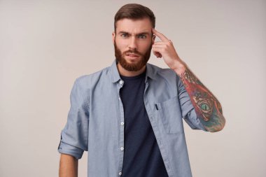 Şakağına parmak büken ve kameraya çapraz bakan, kaşlarını çatarak beyaz arka planda poz veren ciddi yakışıklı ve sakallı bir erkek.