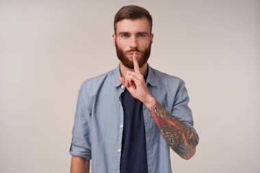 Beyaz arka planda poz verirken mavi tişörtlü, tıraş olmamış, dudaklarında işaret parmağı tutan, ciddi bir yüzle sessiz kalmak isteyen hoş bir genç adamın stüdyo fotoğrafı.