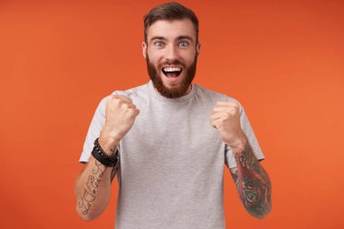 Aşırı mutlu, sakallı ve dövmeli genç bir adam. Heyecanlı yüzüyle kameraya bakıyor ve yumruklarını kaldırıyor. Keyfi yerinde, turuncu arka planda günlük kıyafetler giyiyor.