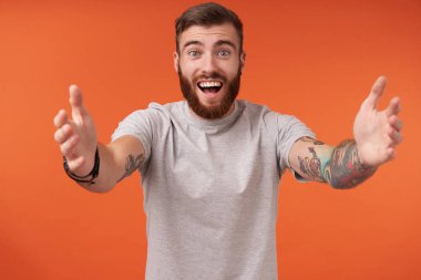 Dövmeli, mutlu ve sakallı bir adam birini gördüğüne seviniyor ve turuncu arka planda dururken bej tişört ve moda aksesuarlar giyerek sarılıyor.