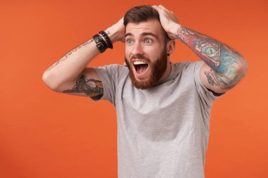 Aşırı neşeli, tıraşsız, esmer, bej tişört ve moda aksesuarlar giyen, başını kaldırmış, şaşırmış yüzüyle yan yana duran, turuncu arka planda izole edilmiş bir adam.