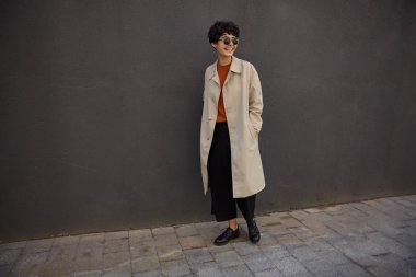 Sıcak bir sonbahar gününde şehir caddesinde yürüyen neşeli, şık, kıvırcık saçlı genç bir kadın, neşeli bir şekilde yan yana bakıyor ve geniş bir şekilde gülümsüyor, sonra siyah beton duvar pozu veriyor.