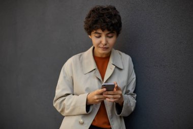 Siyah beton duvarın üzerinde dikilen ve akıllı telefonuyla sosyal ağları kontrol eden, sakin bir yüz ifadesiyle ekrana bakan güzel, siyah saçlı kıvırcık kadın.