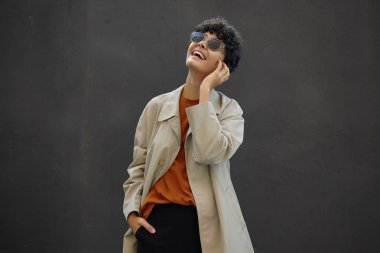 Şehre moda kıyafetler ve güneş gözlükleriyle poz veren mutlu, genç, siyah saçlı kıvırcık kadın mutlu bir şekilde gülüyor ve kafasını geriye atıyor.