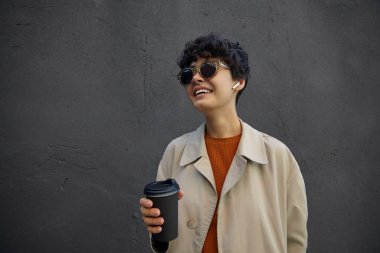 Pozitif, oldukça koyu saçlı, kısa kıvırcık saçlı, modaya uygun kıyafetler giyen ve sokakta yürürken şık güneş gözlüğü takan, işe başlamadan önce sıcak kahve içen bir kadın.