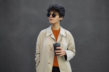 Yaşam tarzı moda portresi. Kısa siyah kıvırcık saçlı, şık kıyafetler ve moda güneş gözlüğü takan, elinde kahve ile caddede yürüyen genç ve şık bir hippi kadın.