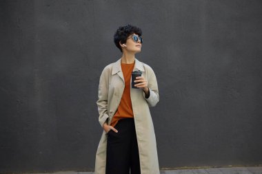 Güzel görünümlü, kıvırcık saçlı, esmer bir bayan, dışarıda kahve içerken, şehir ortamının başında, modaya uygun kıyafetler ve güneş gözlüğü takıyor.