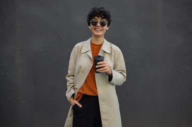 Şaşkın, genç, kıvırcık saçlı, kısa saçlı, elini cebine atmış, kaşlarını kaldırmış kameraya bakan, moda kıyafetlerle şehir ortamının başında duran esmer kadın.