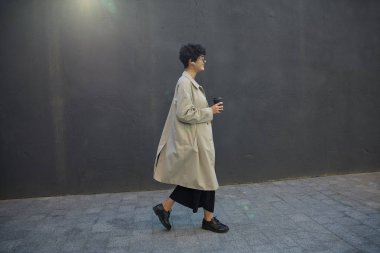 Kısa saçlı, güzel, kıvırcık bir kadının elinde bir fincan kahveyle kentsel beton duvar boyunca yürüdüğü, mutlu ve geniş bir gülümsemeyle ileriye baktığı, güneşli bir kare.