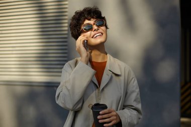 Kısa saçlı, güzel, gülümseyen esmer bir kadının yemek molasında sokakta yürüdüğü ve kahve içtiği, cep telefonuyla telefonla konuştuğu ve mutlu bir şekilde yukarı baktığı bir kare.
