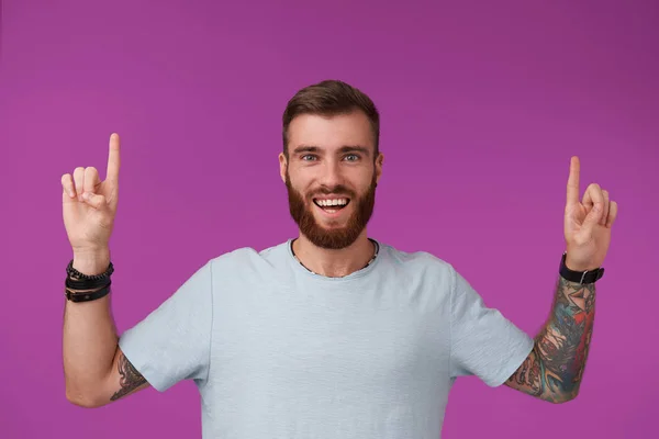 Joven alegre sin afeitar hombre tatuado con corte de pelo corto con camiseta azul mientras está de pie sobre el fondo púrpura, mirando a la cámara con una amplia sonrisa feliz y levantando los dedos índice —  Fotos de Stock