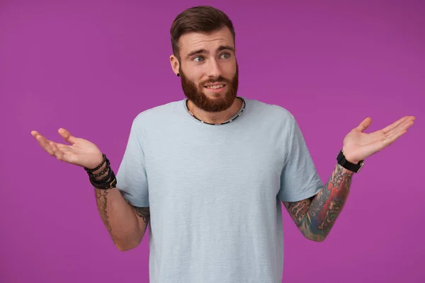 Studio photo of pretty bearded brunette with tattooes looking aside and wrinkling his forehead, posing over purple background and raising palms confusedly — Stock Photo, Image