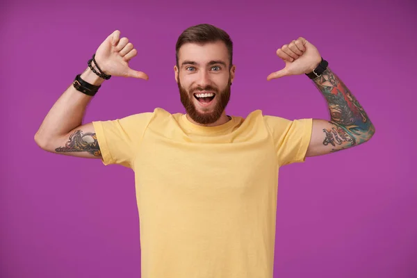 Attractive young brunette bearded male with tattooes raising hands and showing on himself with thumbs, smiling widely to camera with self-confidence, standing over purple background in casual clothes — Stock Photo, Image