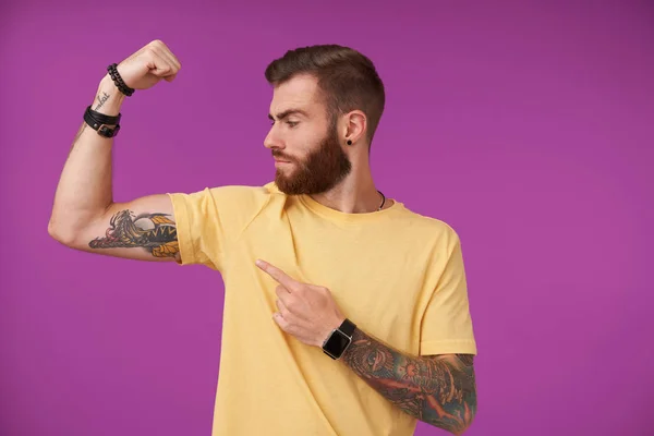 Strong attractive young tattooed male with beard and trendy haircut looking proudly at his hand and demostrating his biceps, standing over purple background in casual t-shirt — Stock Photo, Image