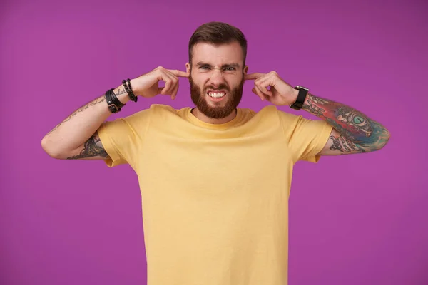 Displeased handsome young tattooed man with beard frowning his face and inserting forefingers to his ears, trying to avoid annoying sounds, isolated over purple background — Stock Photo, Image