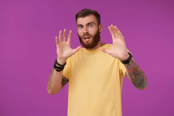 Pop-eyed tattooed brunette unshaved male with short haircut looking scaredly to camera and raising hands to protect himself, standing over purple background in yellow t-shirt — Stock Photo, Image