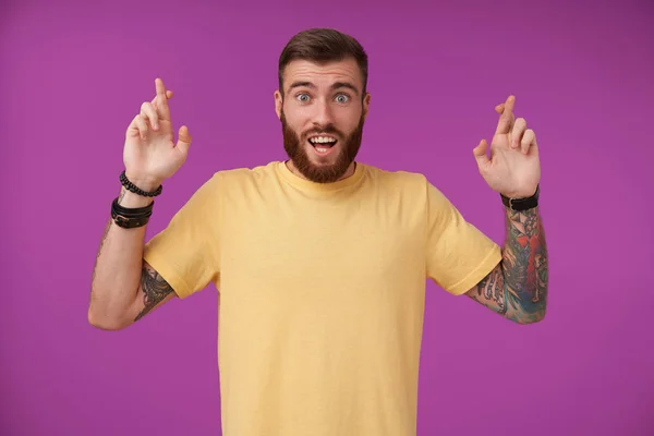Joyful blue-eyed brunette guy with beard and tattooes looking at camera with wide eyes and mouth opened, raising hands with crossed fingers for good luck, isolated over purple background — Stock Photo, Image