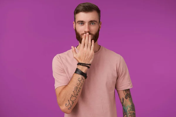 Γοητευτικός νεαρός με τατουάζ γαλανομάτης άνδρας με τατουάζ που φοράει μπεζ t-shirt και μοντέρνα αξεσουάρ ενώ ποζάρει πάνω από μωβ φόντο, κρατώντας την παλάμη στο στόμα του και κοιτάζοντας θετικά την κάμερα — Φωτογραφία Αρχείου