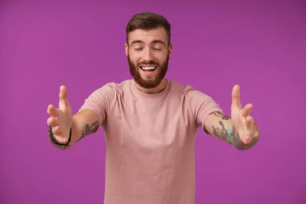 Pleasant looking happy brunette bearded male with tattooes standing over purple background with opened arms, going to give hug and keeping eyes closed — Φωτογραφία Αρχείου