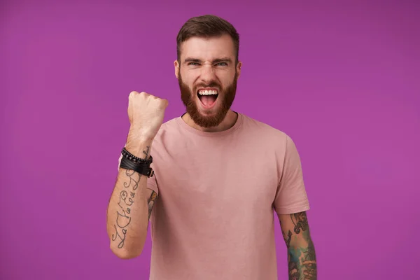 Excited bearded brunette guy with tattooes shouting happily and raising fist in yes sign, frowning face with wide mouth opened, posing over purple background in casual clothes and trendy accessories — Stock Photo, Image