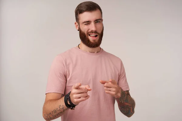 Portrait of young self-confident brunette male with beard wearing beige t-shirt and trendy accessories while posing over white background, winking at camera and keeping index fingers raised