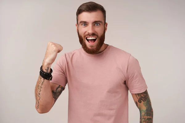 Good looking glad young tattooed brunette man with trendy haircut smiling widely and raising fist in yes gesture, wearing beige t-shirt and trendy accessories while standing over white background — Stock Photo, Image