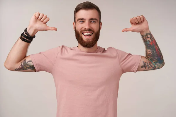 Happy attractive young bearded male with tattooes and short haircut wearing casual clothes while posing over white background, looking joyfully to camera and pointing to himself with raised thumbs — Stock Photo, Image