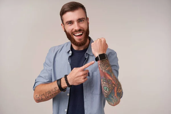 Joyful young pretty bearded brunette man with tattooed hands smiling widely to camera and pointing to his watch, being in high spirit while posing over white background — Stock Photo, Image