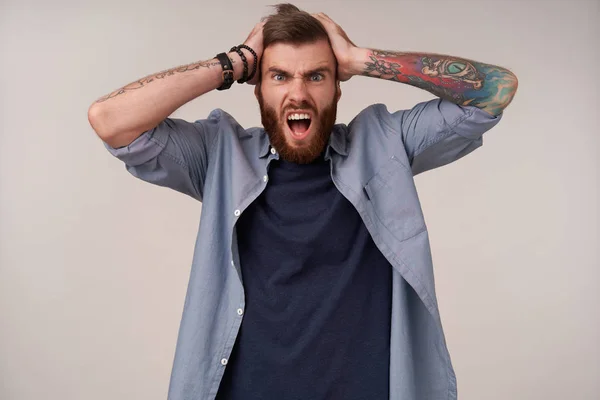 Stressed handsome young blue-eyed man with tattooes wearing blue t-shirt while posing over white background, looking at camera and screaming, holding his head with raised hands — Stock Photo, Image