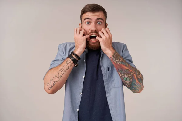 Εσωτερική φωτογραφία ενός φοβισμένου μελαχρινού με τατουάζ με γενειάδα που κοιτάζει την κάμερα με ανοιχτά μάτια και το στόμα ανοιχτό, κρατώντας τα χέρια στο πρόσωπό του ενώ στέκεται πάνω από το λευκό φόντο — Φωτογραφία Αρχείου