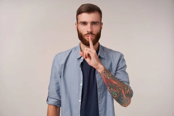 Στιγμιότυπο ενός όμορφου νεαρού αξύριστου άντρα με τατουάζ που φοράει μπλε πουκάμισο ενώ ποζάρει πάνω από λευκό φόντο, κρατώντας δείκτη στα χείλη του, ζητώντας να κρατήσει τη σιωπή με σοβαρό πρόσωπο — Φωτογραφία Αρχείου