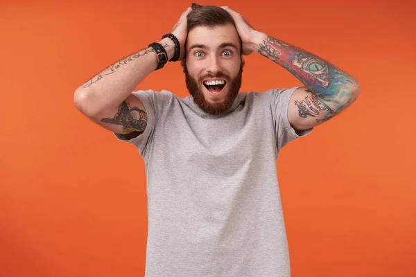 Overjoyed pretty bearded brunette man with tattooes clutching his head with raised hands and looking happily to camera, standing over orange background in casual clothes — Stock Photo, Image