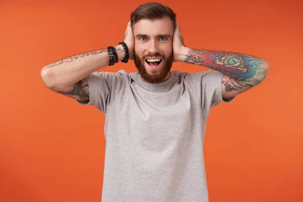 Χαρούμενος γενειοφόρος μελαχρινός άντρας με τατουάζ που καλύπτει τα αυτιά του με υψωμένες παλάμες και κοιτάζει χαρούμενα την κάμερα, αποδομώντας τα λευκά τέλεια δόντια του ενώ στέκεται πάνω από πορτοκαλί φόντο — Φωτογραφία Αρχείου