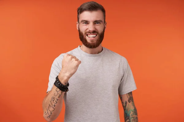 Pleased young tattooed brunette male with beard wearing beige t-shirt and trendy accessories while standing over orange background, raising fist happily in yes gesture — Stock Photo, Image