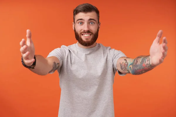 Studio shot of positive pretty young tattooed male with lush beard posing over orange background with opened arms, going to give hug, showing his pleasant emotions — Stock Photo, Image