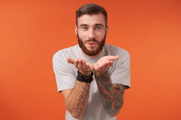 stock image Positive young tattooed brunette male with lush beard looking romantically to camera and sending air kiss to camera, keeping hands raised while posing over orange background