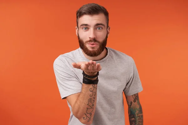 Charming lovely young bearded brunette guy with short haircut raising his hand and blowing positively air kiss to camera, standing over orange background in casual clothes — Stock Photo, Image