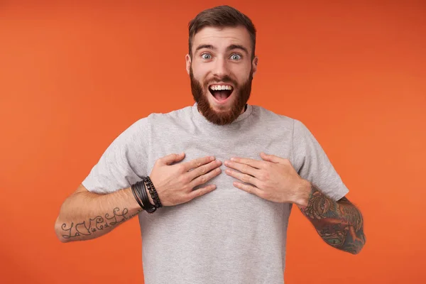 Overjoyed pretty young bearded brunette male with trendy haircut looking at camera happily and rounding eyes, keeping palms on his chest while standing over orange background — Stock Photo, Image