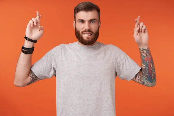 Puzzled blue-eyed young bearded man with trendy haircut raising hands with crossed fingers, hoping for better, frowning eyebrows and biting underlip while standing over orange background — Stock Photo, Image