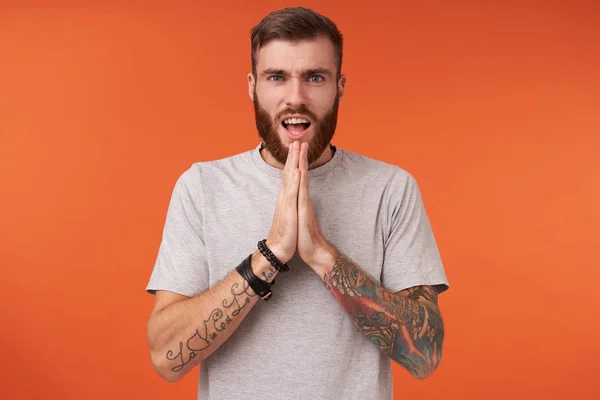 Indoor photo of serious young bearded brunette male in beige t-shirt and trendy accessories posing over orange background with folded hands, frowning eyebrows and opening mouth — Stock Photo, Image