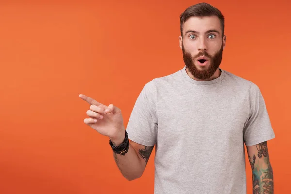 Suprised bearded blue-eyed man with tattooes showing aside with index finger and rounding eyes while looking at camera, wearing beige t-shirt while posing over orange background