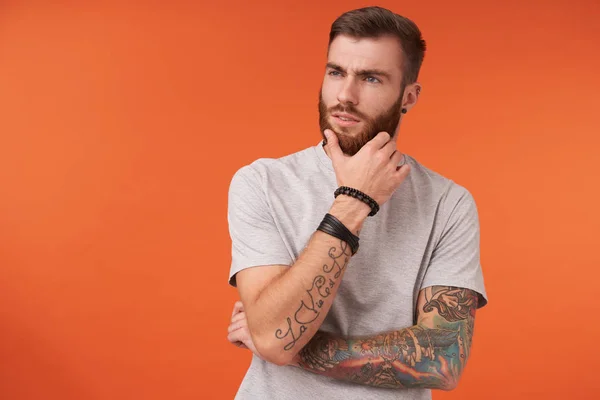 Pensive νεαρός μελαχροινή με τατουάζ άνδρας σε μπεζ t-shirt και μοντέρνα αξεσουάρ κρατώντας το χέρι στην πλούσια γενειάδα του και κοιτάζοντας πέρα προσεκτικά, απομονώνονται πάνω από πορτοκαλί φόντο — Φωτογραφία Αρχείου