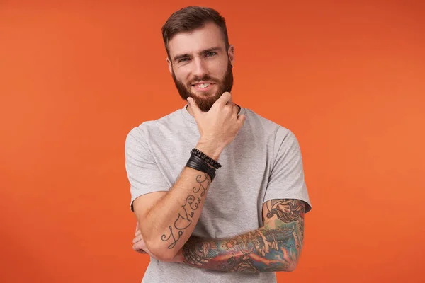 Νεαρός ελκυστικός γενειοφόρος άνδρας με τατουάζ και μοντέρνο κούρεμα κοιτάζοντας την κάμερα με γοητευτικό χαμόγελο και κρατώντας το πηγούνι του με υψωμένο χέρι, ποζάροντας πάνω από πορτοκαλί φόντο σε casual wear — Φωτογραφία Αρχείου