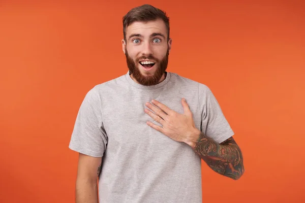 Studio shot of blue-eyed unshaved tattooed man keeping palm on his chest and looking to camera with surprised face, raising eyebrows and wrinkling forehead over orange background — Stock Photo, Image